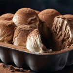 Overhead shot of creamy Tiramisu Ice Cream in a pan with cocoa powder dusting and a scoop removed.1